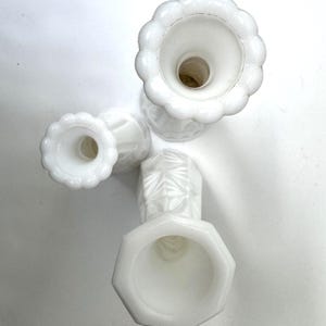May include: Four white, vintage-style candle holders. The candle holders have different shapes, including a scalloped edge and an octagonal base. The candle holders are made of a milky white glass and are arranged on a white surface.