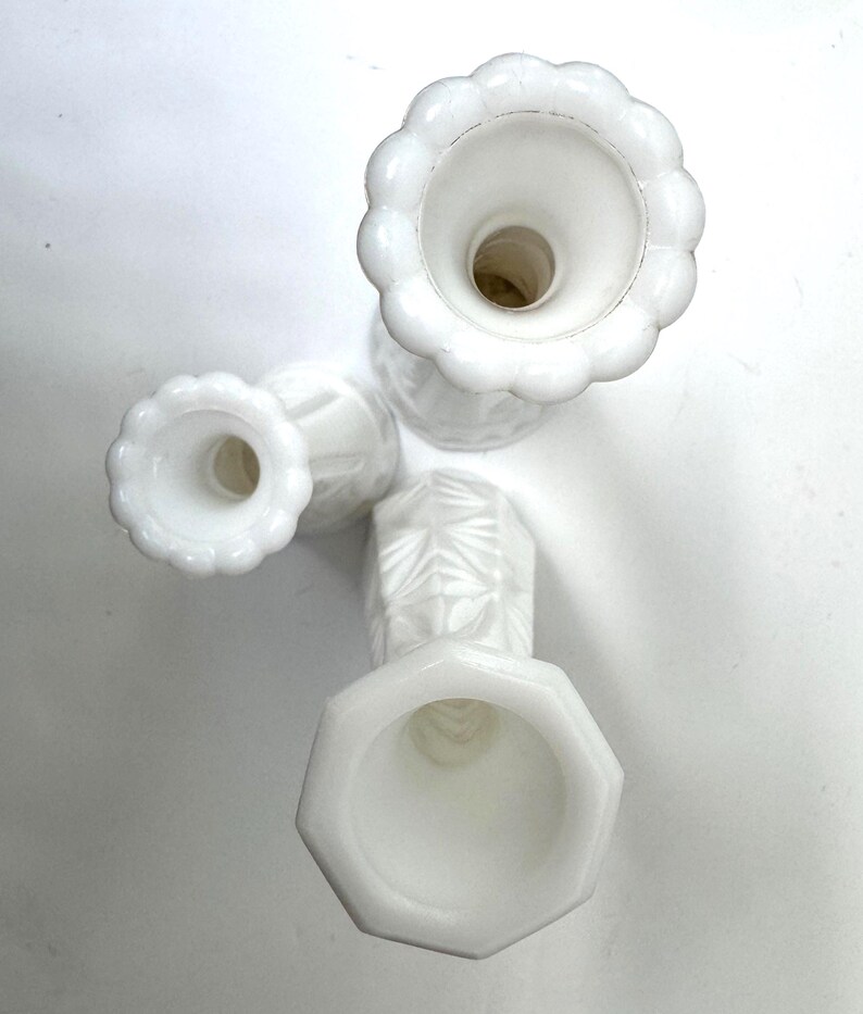May include: Four white, vintage-style candle holders. The candle holders have different shapes, including a scalloped edge and an octagonal base. The candle holders are made of a milky white glass and are arranged on a white surface.