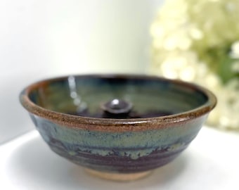 May include: A handmade ceramic bowl with a dark interior and a rim with brown and blue-green hues. The bowl has a small, dark, round object in the center. The exterior has a textured, layered appearance.
