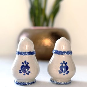 Può includere: Un paio di saliere e pepiere in ceramica. I contenitori sono bianco sporco con disegni a cuore floreale blu. Rifiniture blu decorano la parte superiore e inferiore di ogni contenitore. Lo sfondo è sfocato, con un vaso dorato e una pianta verde.