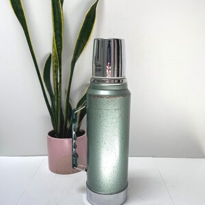 May include: A vintage-style sage green vacuum insulated thermos with a metallic silver cap and base. The cylindrical body features a textured finish with visible signs of wear near the top and a dark green plastic handle attached to the side.