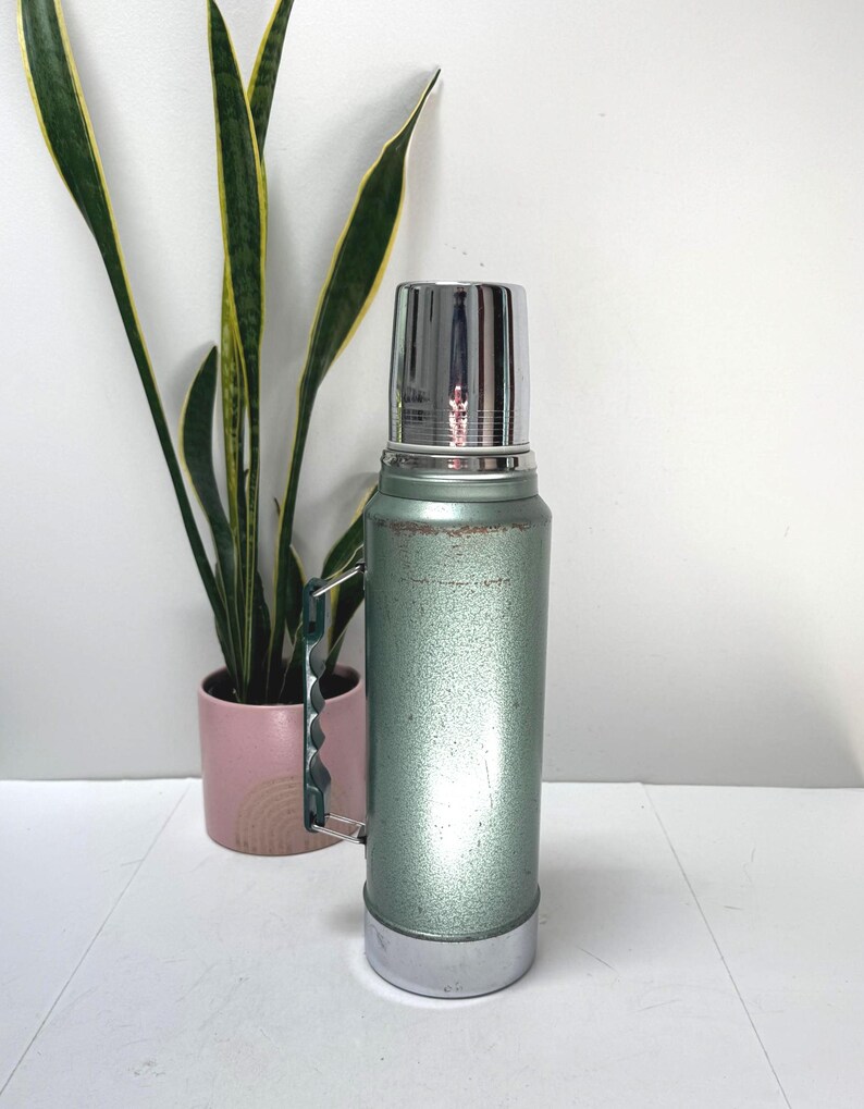 May include: A vintage-style sage green vacuum insulated thermos with a metallic silver cap and base. The cylindrical body features a textured finish with visible signs of wear near the top and a dark green plastic handle attached to the side.
