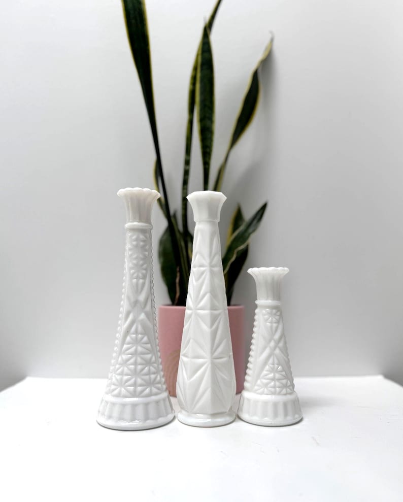 May include: Three white, decorative vases of varying heights. The vases have textured geometric patterns and flared tops. A potted plant with green and yellow leaves is in the background, adding a touch of natural color to the composition.