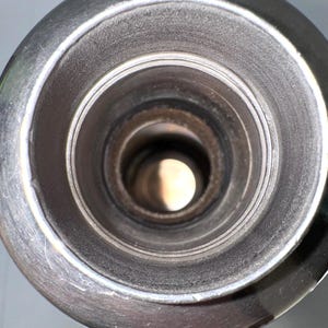 May include: A top-down view of a cylindrical metal container opening, featuring a wide, circular rim with a smooth, polished finish. The interior walls are dark and textured, leading down to a deep, hollow center that reveals a bright light.