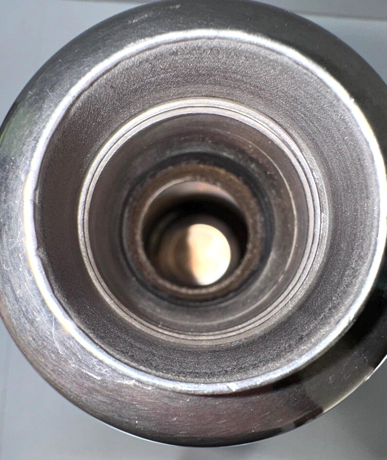 May include: A top-down view of a cylindrical metal container opening, featuring a wide, circular rim with a smooth, polished finish. The interior walls are dark and textured, leading down to a deep, hollow center that reveals a bright light.