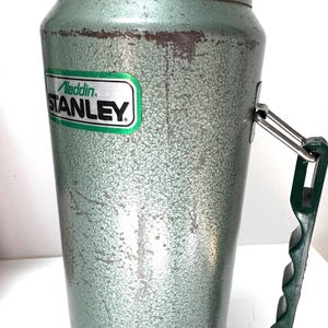 May include: A vintage green metal vacuum flask with a textured hammer-tone finish. The body features a rectangular green and silver label with the text Aladdin STANLEY. A dark green plastic handle is attached to the side with a metal hinge.