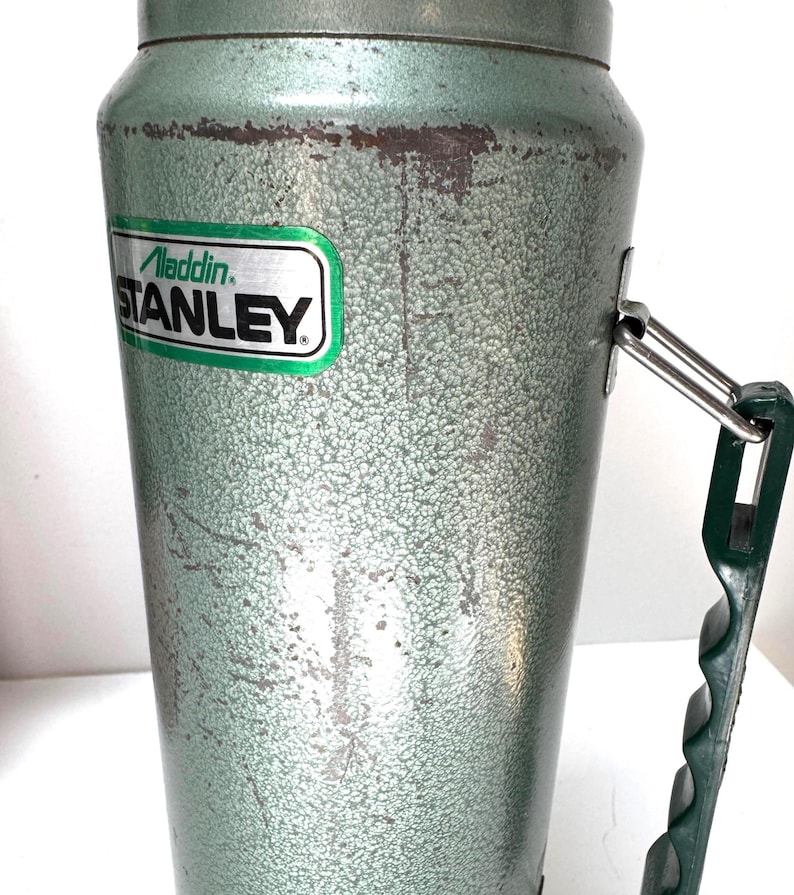 May include: A vintage green metal vacuum flask with a textured hammer-tone finish. The body features a rectangular green and silver label with the text Aladdin STANLEY. A dark green plastic handle is attached to the side with a metal hinge.