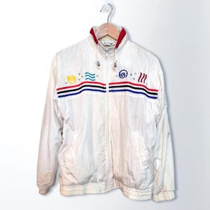 May include: A white windbreaker jacket featuring a full-length front zipper, a high collar with red trim, and long sleeves. The chest displays horizontal black, red, and blue stripes with embroidered yellow, blue, and red abstract symbols above.