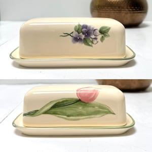 May include: A cream-colored ceramic butter dish with a rectangular base and a matching lid. The lid features two distinct floral designs: a cluster of purple flowers with green leaves on one side and a single pink tulip with green leaves on the other.