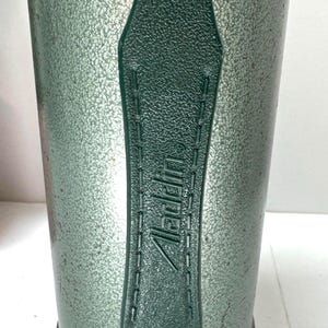 May include: A vintage green vacuum flask with a textured, hammered metal finish and a dark green plastic handle. The handle features the embossed text Aladdin with a registered trademark symbol. The base of the flask is made of polished silver metal.