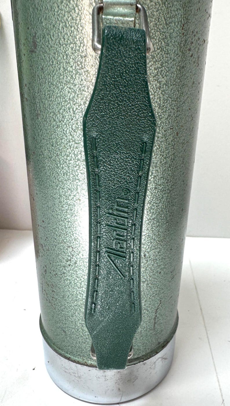 May include: A vintage green vacuum flask with a textured, hammered metal finish and a dark green plastic handle. The handle features the embossed text Aladdin with a registered trademark symbol. The base of the flask is made of polished silver metal.