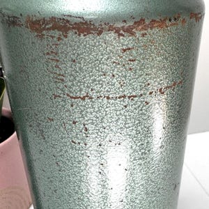 May include: A cylindrical metal canister with a textured, sage green hammered finish. The surface displays visible signs of wear, including horizontal bands of brown rust and scattered scratches across the exterior body of the container.