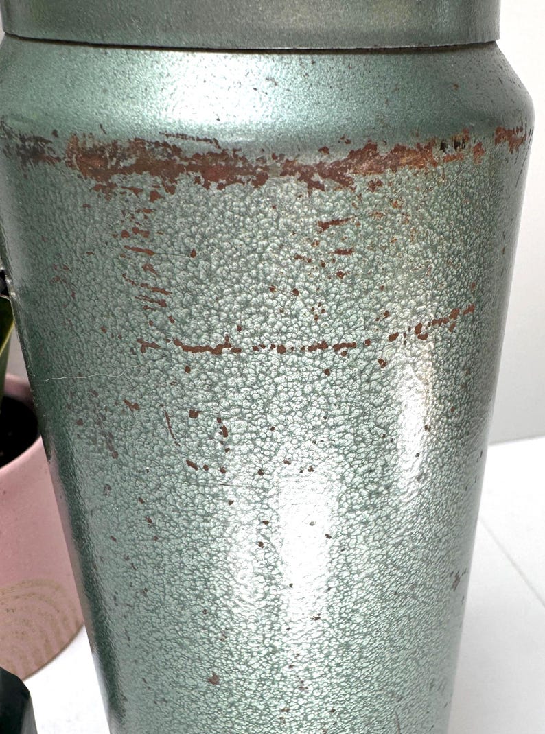 May include: A cylindrical metal canister with a textured, sage green hammered finish. The surface displays visible signs of wear, including horizontal bands of brown rust and scattered scratches across the exterior body of the container.