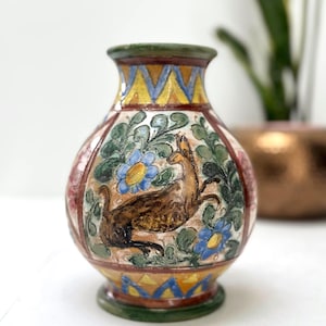 Könnte beinhalten: Kleine, runde Keramikvase mit detailliertem, gemaltem Design. Die Vase zeigt einen braunen Hirsch, blaue Blumen und grünes Laub. Der Rand und der Boden sind grün, mit gelben und blauen geometrischen Mustern. Die Vase ist etwa 15 cm hoch.
