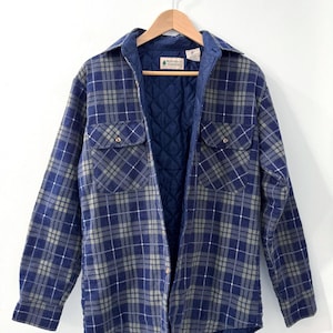 May include: A blue and grey checked flannel shirt jacket with a quilted navy lining. The shirt has two chest pockets and a button-down front. The jacket is hanging on a wooden hanger.