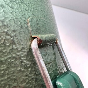 May include: A sage green container with a textured, hammered finish featuring a silver metal latch mechanism. The metal hinge shows visible signs of oxidation and rust where it connects to the green body of the container.