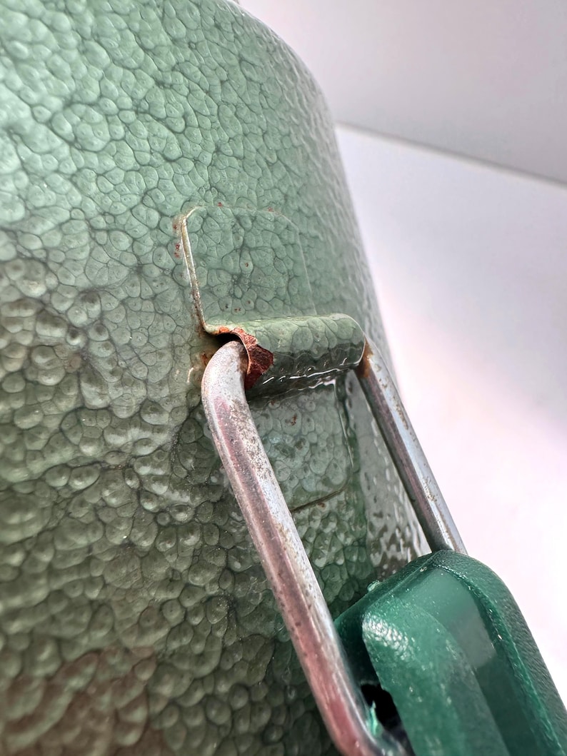 May include: A sage green container with a textured, hammered finish featuring a silver metal latch mechanism. The metal hinge shows visible signs of oxidation and rust where it connects to the green body of the container.