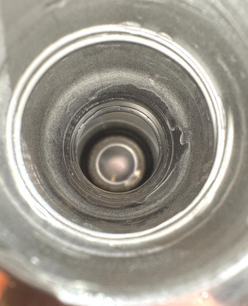 May include: A top-down view into the cylindrical opening of a metallic bottle. The interior walls are smooth and silver-toned, leading down to a circular base. The rim of the opening features a threaded neck design for a cap, with a metallic finish.