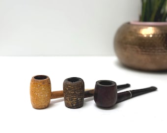 May include: Three tobacco pipes of varying designs are arranged on a white surface. The first pipe is made of light-colored corn cob, the second is dark brown, and the third is a dark wood with a black stem. A decorative pot is in the background.