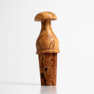 May include: A bottle stopper featuring a smooth, light brown wooden top with a rounded mushroom-like shape and a distinct grain pattern, attached to a tapered, porous, light brown cork base with a textured, speckled surface appearance.