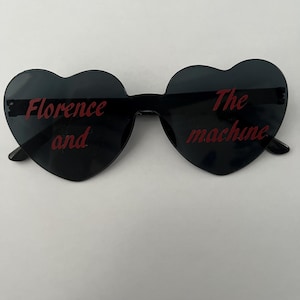 May include: Black heart-shaped sunglasses with the text "Florence and the Machine" in red. The sunglasses have a dark tint and a black frame. The text is in a handwritten style.