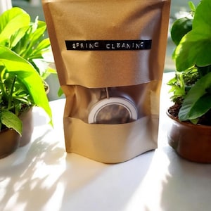 Może przedstawiać: Brązowa papierowa torba z napisem "SPRING CLEANING" na czarnej etykiecie. Torba jest częściowo otwarta, odsłaniając pojemnik ze srebrną pokrywką. Zielone rośliny są widoczne w tle, tworząc naturalną estetykę.