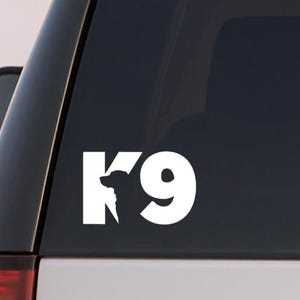 May include: White K9 decal on a vehicle's rear window. The decal features the letters "K9" with a silhouette of a dog's head incorporated into the "K". The image is a close-up, showing the decal's clean lines and placement on the car.