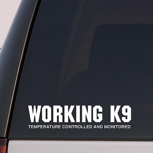 May include: White decal on a vehicle's rear window. The decal reads "WORKING K9" in bold, capitalized letters, with "TEMPERATURE CONTROLLED AND MONITORED" below in smaller text. The vehicle is white.