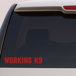 Peut inclure: Autocollant en vinyle rouge avec l'inscription "WORKING K9" sur la vitre arrière d'un véhicule. Le texte est en gras, en police sans-serif. Le véhicule est blanc avec une vitre teintée foncée.