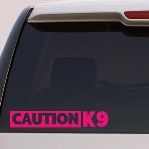 May include: A pink sticker on a car window, displaying the words "CAUTION K9" in a bold, sans-serif font. The sticker is a vibrant pink and is positioned horizontally on the rear window of a white vehicle.