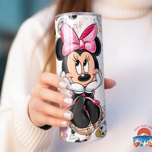 May include: A white tumbler featuring a Minnie Mouse design. The tumbler has a pink bow, black ears, and a plaid skirt. Floral and character accents surround the character. The tumbler is cylindrical and appears to be made of stainless steel.