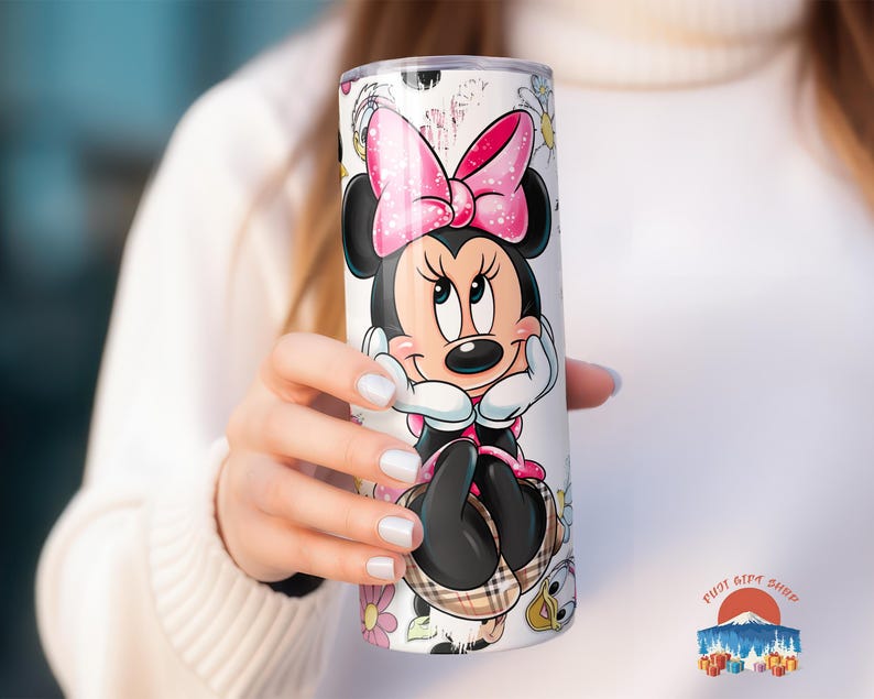 May include: A white tumbler featuring a Minnie Mouse design. The tumbler has a pink bow, black ears, and a plaid skirt. Floral and character accents surround the character. The tumbler is cylindrical and appears to be made of stainless steel.