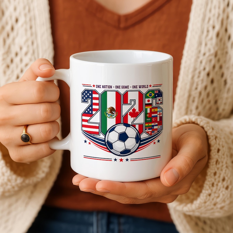 Puede incluir: Una taza de cer&aacute;mica blanca con el texto 2026, donde los n&uacute;meros est&aacute;n rellenos con las banderas de Estados Unidos, M&eacute;xico y Canad&aacute;, junto a un gr&aacute;fico de un bal&oacute;n de f&uacute;tbol y el texto ONE NATION - ONE GAME - ONE WORLD en azul y rojo.