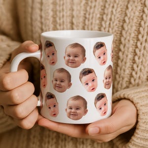 May include: White ceramic mug featuring multiple baby faces. The faces are of various babies with different expressions and hair colors. The mug has a white handle and is held by a person wearing a beige sweater.