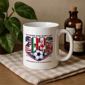 Puede incluir: Una taza de cer&aacute;mica blanca con el texto 'ONE NATION - ONE GAME - ONE WORLD' sobre el a&ntilde;o '2026'. Los n&uacute;meros est&aacute;n rellenos con varias banderas nacionales, incluidas las de EE. UU., M&eacute;xico y Canad&aacute;, con un gr&aacute;fico de un bal&oacute;n de f&uacute;tbol.