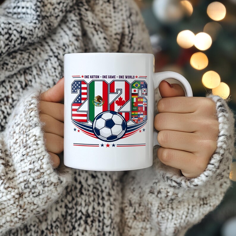Puede incluir: Una taza de cer&aacute;mica blanca con el texto 2026, cuyos n&uacute;meros est&aacute;n rellenos con banderas nacionales, incluyendo las de EE. UU., M&eacute;xico y Canad&aacute;. Debajo del texto hay un gr&aacute;fico de un bal&oacute;n de f&uacute;tbol y la frase ONE NATION ONE GAME ONE WORLD.