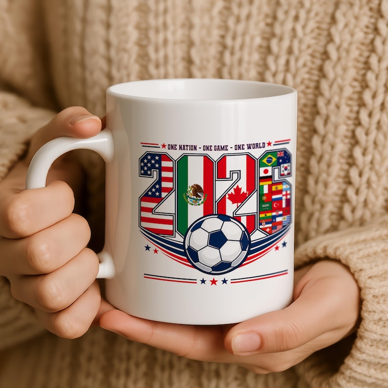 Puede incluir: Una taza de cer&aacute;mica blanca con el texto 'ONE NATION - ONE GAME - ONE WORLD' sobre el a&ntilde;o '2026'. Los n&uacute;meros est&aacute;n rellenos con varias banderas nacionales, incluyendo las de EE. UU., M&eacute;xico y Canad&aacute;, y un bal&oacute;n de f&uacute;tbol debajo del texto.