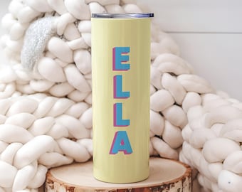 May include: A light yellow tumbler with the name "ELLA" in blue letters with a pink outline. The tumbler is cylindrical and sits on a wooden slice. The background features a chunky, off-white knitted blanket.