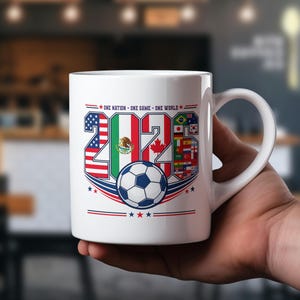 Puede incluir: Una taza de cer&aacute;mica blanca con un dise&ntilde;o gr&aacute;fico del a&ntilde;o 2026, con los n&uacute;meros rellenos con las banderas de Estados Unidos, M&eacute;xico y Canad&aacute;, junto a un bal&oacute;n de f&uacute;tbol y el texto One Nation - One Game - One World impreso en la parte superior.
