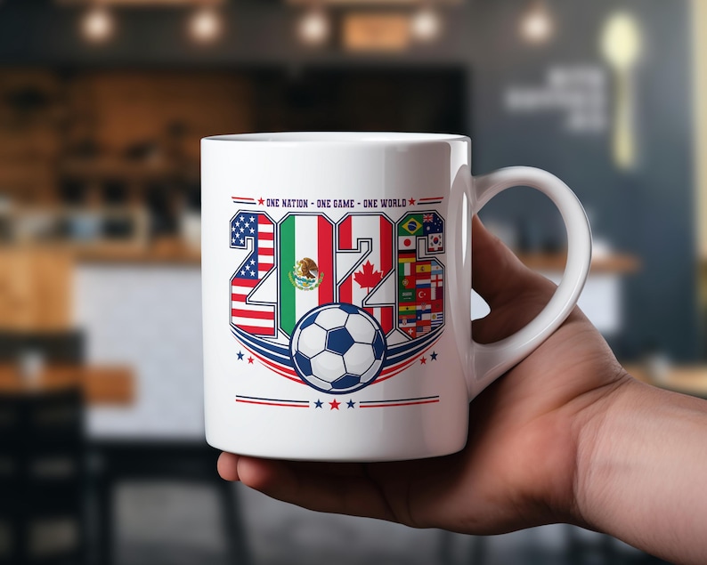 Puede incluir: Una taza de cer&aacute;mica blanca con un dise&ntilde;o gr&aacute;fico del a&ntilde;o 2026, con los n&uacute;meros rellenos con las banderas de Estados Unidos, M&eacute;xico y Canad&aacute;, junto a un bal&oacute;n de f&uacute;tbol y el texto One Nation - One Game - One World impreso en la parte superior.