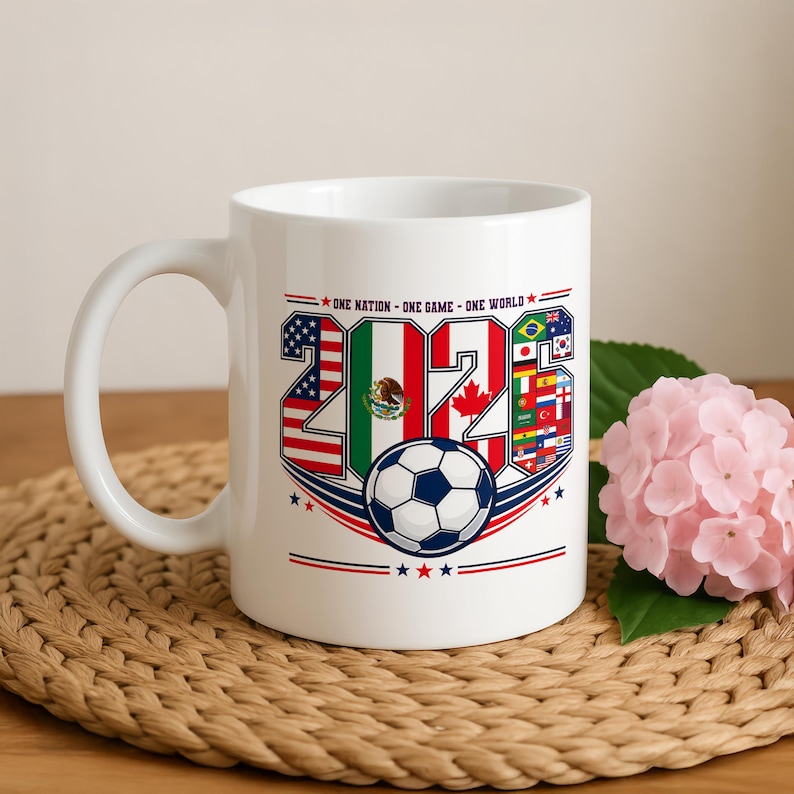 Puede incluir: Una taza de cer&aacute;mica blanca con el texto 'ONE NATION - ONE GAME - ONE WORLD' sobre el a&ntilde;o '2026'. Los n&uacute;meros est&aacute;n rellenos con varias banderas nacionales, incluyendo las de EE. UU., M&eacute;xico y Canad&aacute;, con un gr&aacute;fico de un bal&oacute;n de f&uacute;tbol.