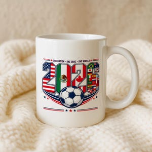 Puede incluir: Una taza de cer&aacute;mica blanca con el texto ONE NATION - ONE GAME - ONE WORLD sobre el a&ntilde;o 2026. Los n&uacute;meros est&aacute;n rellenos con las banderas de Estados Unidos, M&eacute;xico, Canad&aacute; y otras naciones, con un gr&aacute;fico de un bal&oacute;n de f&uacute;tbol debajo.