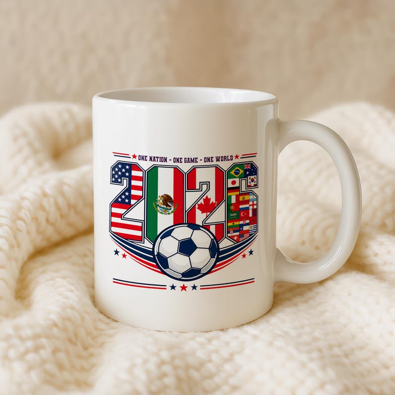 Puede incluir: Una taza de cer&aacute;mica blanca con el texto ONE NATION - ONE GAME - ONE WORLD sobre el a&ntilde;o 2026. Los n&uacute;meros est&aacute;n rellenos con las banderas de Estados Unidos, M&eacute;xico, Canad&aacute; y otras naciones, con un gr&aacute;fico de un bal&oacute;n de f&uacute;tbol debajo.