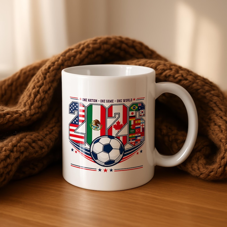 Puede incluir: Una taza de cer&aacute;mica blanca con un dise&ntilde;o gr&aacute;fico que incluye el texto 'ONE NATION - ONE GAME - ONE WORLD' y el a&ntilde;o '2026'. Los n&uacute;meros est&aacute;n rellenos con varias banderas nacionales y un bal&oacute;n de f&uacute;tbol est&aacute; centrado debajo del texto con detalles en rojo y azul.