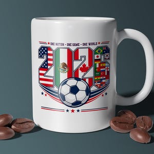 Puede incluir: Una taza de cer&aacute;mica blanca con el texto 2026, cuyos n&uacute;meros est&aacute;n rellenos con las banderas de Estados Unidos, M&eacute;xico y Canad&aacute;. Debajo del texto hay un gr&aacute;fico de un bal&oacute;n de f&uacute;tbol y la frase ONE NATION - ONE GAME - ONE WORLD con detalles en rojo y azul.