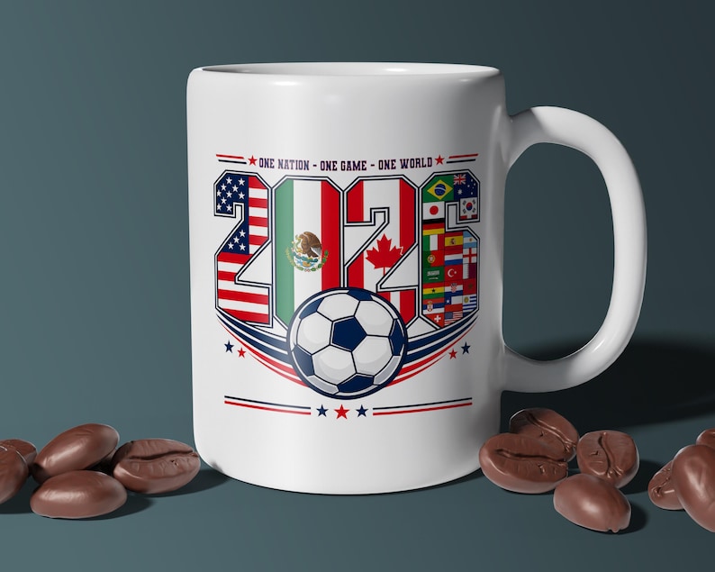 Puede incluir: Una taza de cer&aacute;mica blanca con el texto 2026, cuyos n&uacute;meros est&aacute;n rellenos con las banderas de Estados Unidos, M&eacute;xico y Canad&aacute;. Debajo del texto hay un gr&aacute;fico de un bal&oacute;n de f&uacute;tbol y la frase ONE NATION - ONE GAME - ONE WORLD con detalles en rojo y azul.