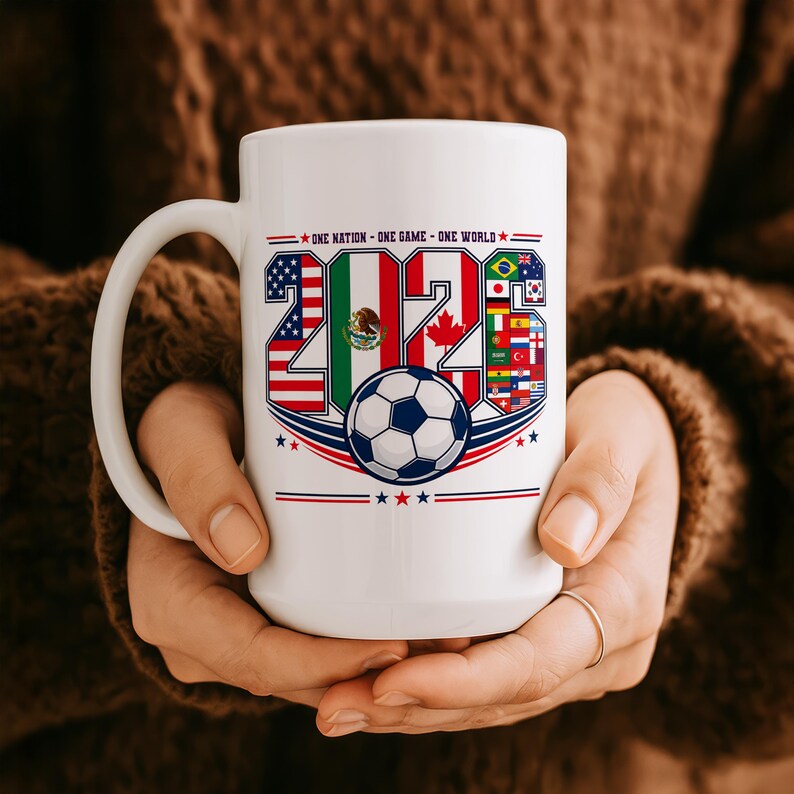 Puede incluir: Una taza de cer&aacute;mica blanca con el texto ONE NATION - ONE GAME - ONE WORLD sobre el a&ntilde;o 2026. Los n&uacute;meros est&aacute;n rellenos con las banderas de EE. UU., M&eacute;xico y Canad&aacute;, junto a un gr&aacute;fico de un bal&oacute;n de f&uacute;tbol y varias banderas internacionales.