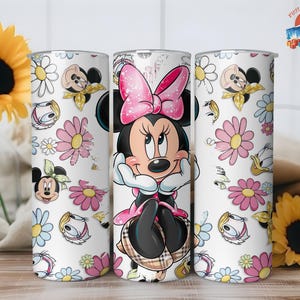 May include: A white tumbler featuring Minnie Mouse in a pink dress and bow, surrounded by cartoon flowers and character faces. The tumbler has a stainless steel appearance and is decorated with a repeating pattern of daisies and other floral elements.