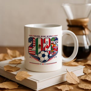 Puede incluir: Una taza de cer&aacute;mica blanca con el texto 2026, cuyos n&uacute;meros est&aacute;n rellenos con varias banderas nacionales, incluyendo las de Estados Unidos, M&eacute;xico y Canad&aacute;. Debajo del texto hay un gr&aacute;fico de un bal&oacute;n de f&uacute;tbol y la frase ONE NATION - ONE GAME - ONE WORLD.