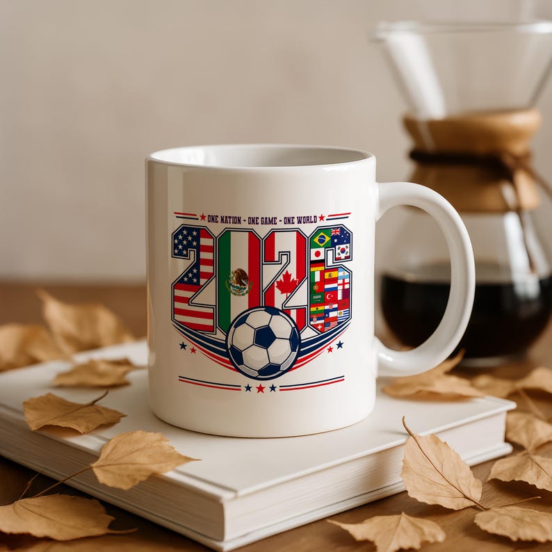 Puede incluir: Una taza de cer&aacute;mica blanca con el texto 2026, cuyos n&uacute;meros est&aacute;n rellenos con varias banderas nacionales, incluyendo las de Estados Unidos, M&eacute;xico y Canad&aacute;. Debajo del texto hay un gr&aacute;fico de un bal&oacute;n de f&uacute;tbol y la frase ONE NATION - ONE GAME - ONE WORLD.